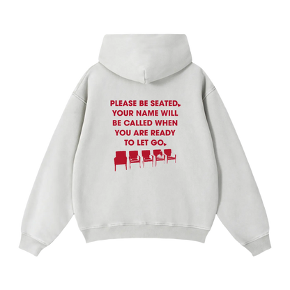 A light gray hoodie shown from the back, featuring a red printed graphic that reads 'PLEASE BE SEATED. YOUR NAME WILL BE CALLED WHEN YOU ARE READY TO LET GO.'