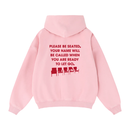 A light pink pullover hoodie shown from the back, featuring a red printed text graphic that reads 'PLEASE BE SEATED. YOUR NAME WILL BE CALLED WHEN YOU ARE READY TO LET GO.'